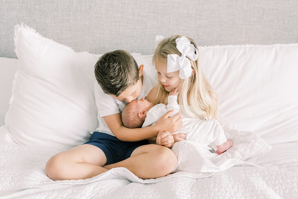 A boy and girl pose for a portrait with their newborn brother while sitting on their bed.
