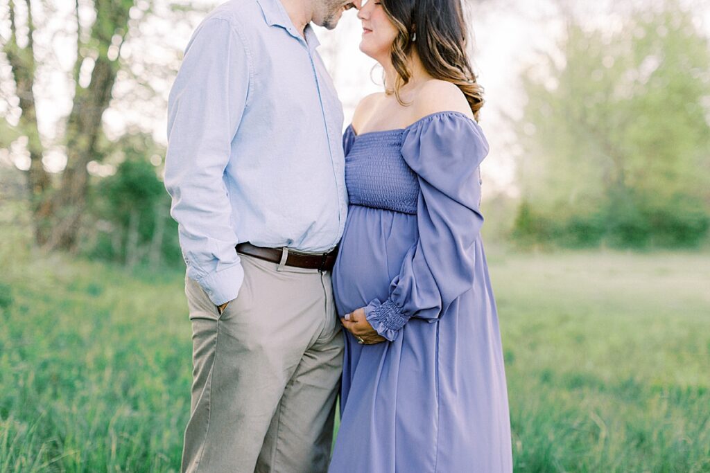 A pregnant woman in a purple dress poses with her husband in a field.
