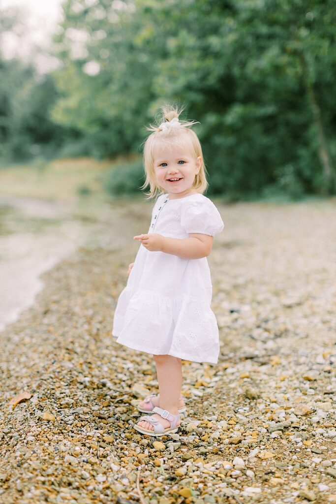 A little girl poses for a portrait next to a lake while wearing a white dress.