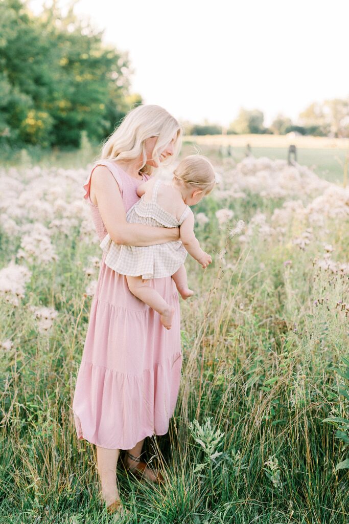 A mother holds her daughter has she reaches for a flower in a field in a photo by Indianapolis family photographer Katelyn Ng Photography.