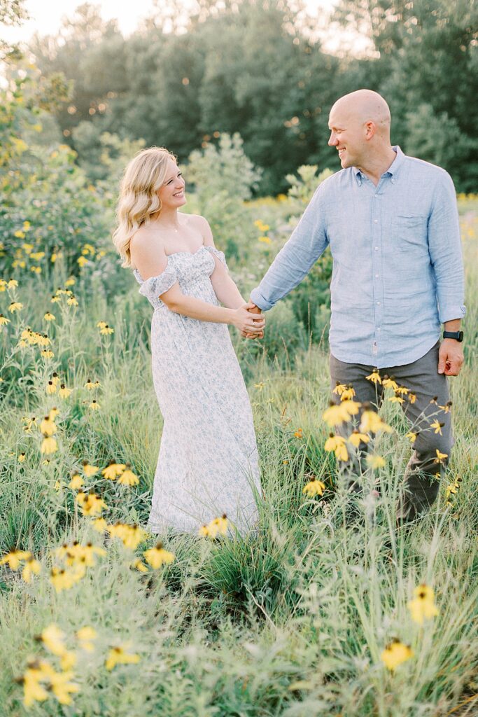 A couple holds hands, surrounded by tall grasses and wildflowers.