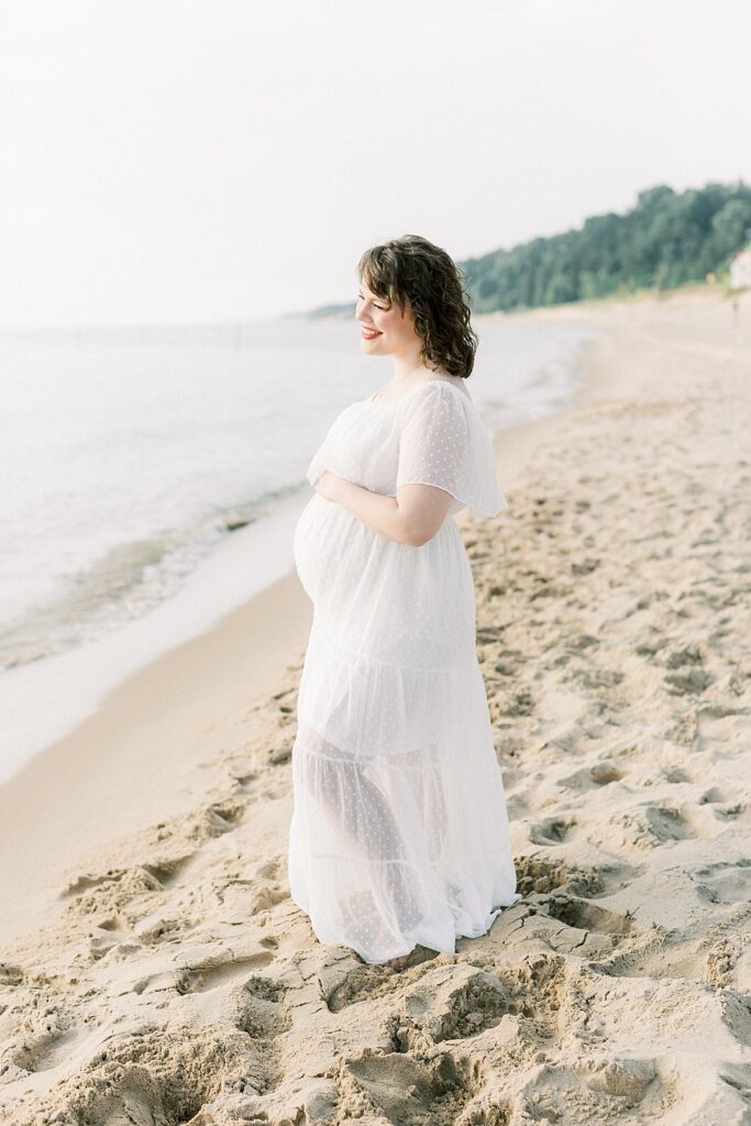 A pregnant woman stands on a beach looking over the water in Holland Michigan in a maternity photo by Katelyn Ng Photography.