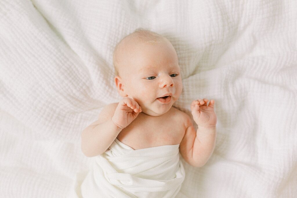 A newborn boy looks around while laying on a white muslin blanket. 