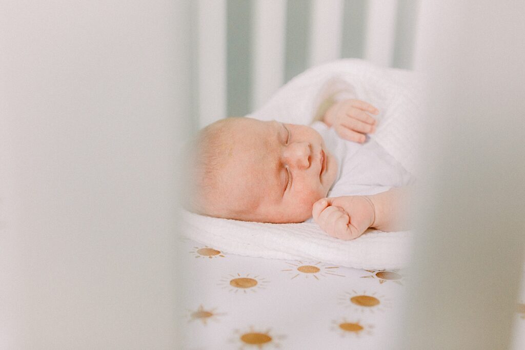 A little boy wrapped in a white blanket sleeps in his crib during his in home newborn photo shoot by Katelyn Ng Photography.  