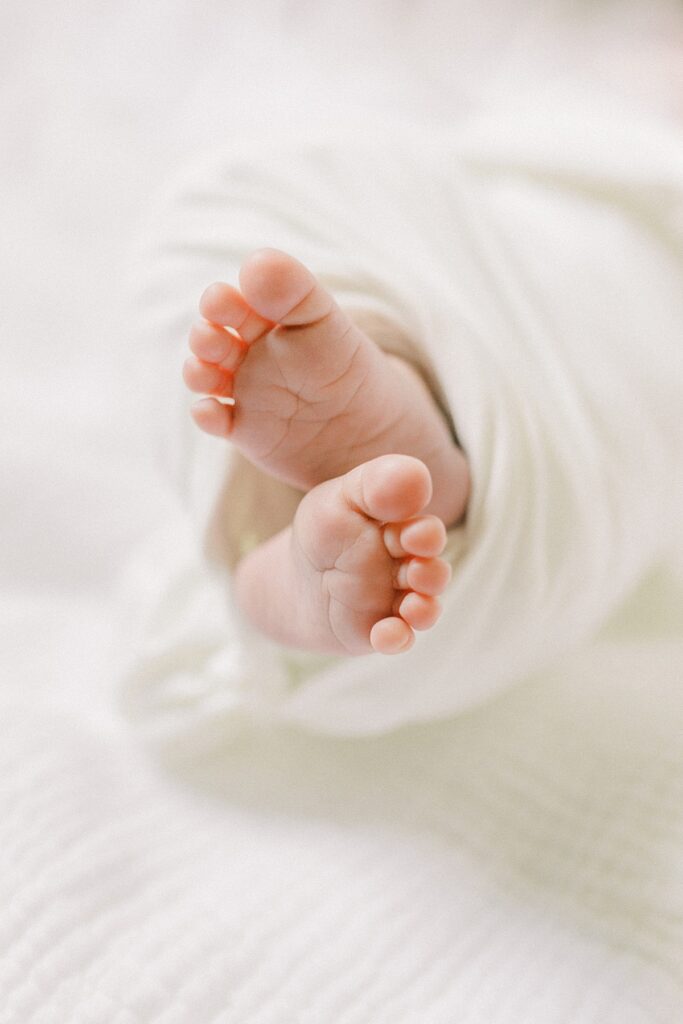 A newborn baby is posed so that his little toes peek out from the white blanket that swaddles him. 