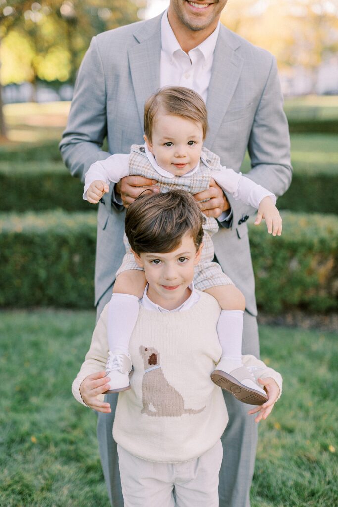 A little boy sits on his brother's shoulders, supported by his father who wears a gray suit coat.