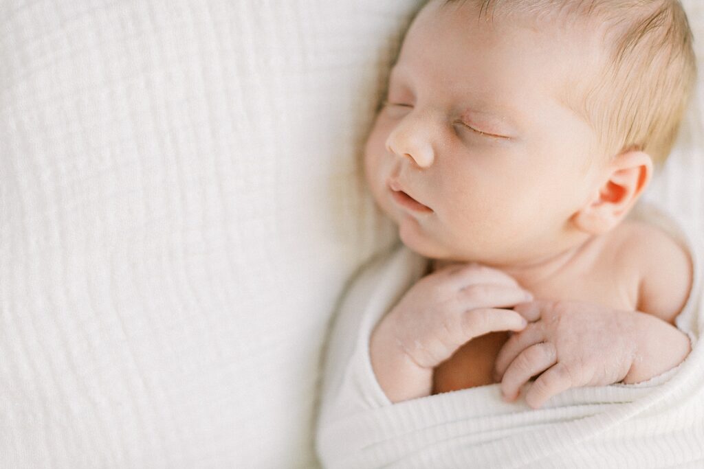 A newborn boy lays swaddled in a white blanket on a muslin sheet.