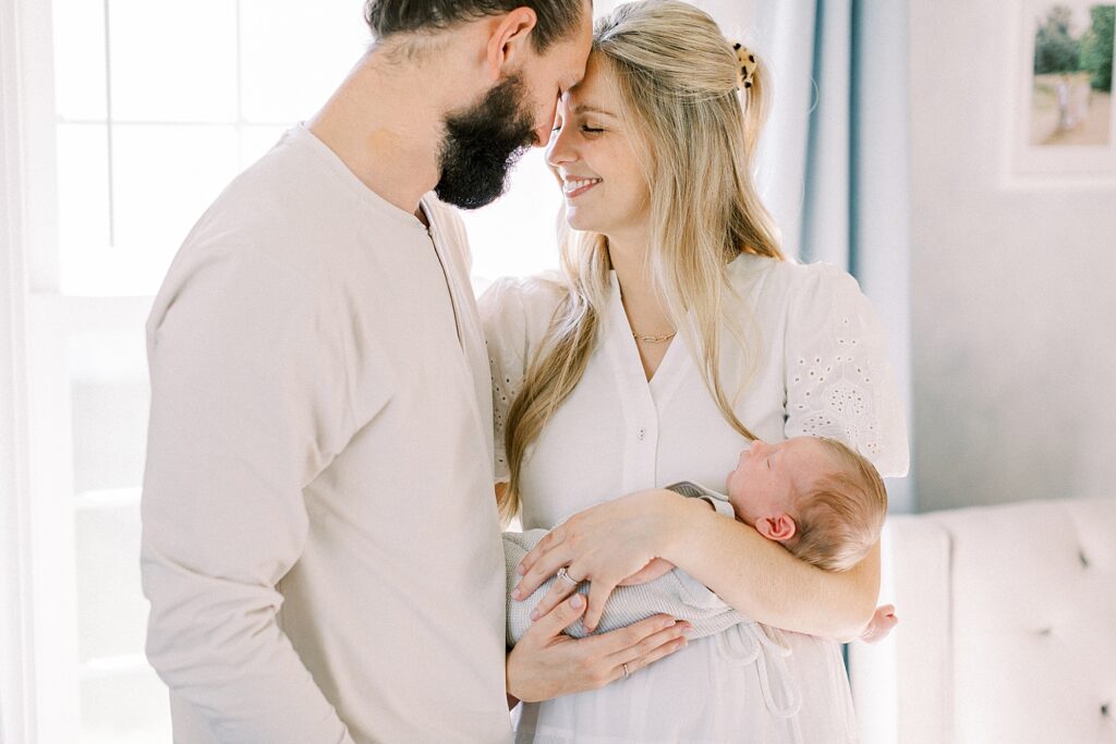 A husband and wife snuggle their newborn baby boy in front of a sunny window in their nursery in a newborn photo by Indianapolis photographer, Katelyn Ng Photography