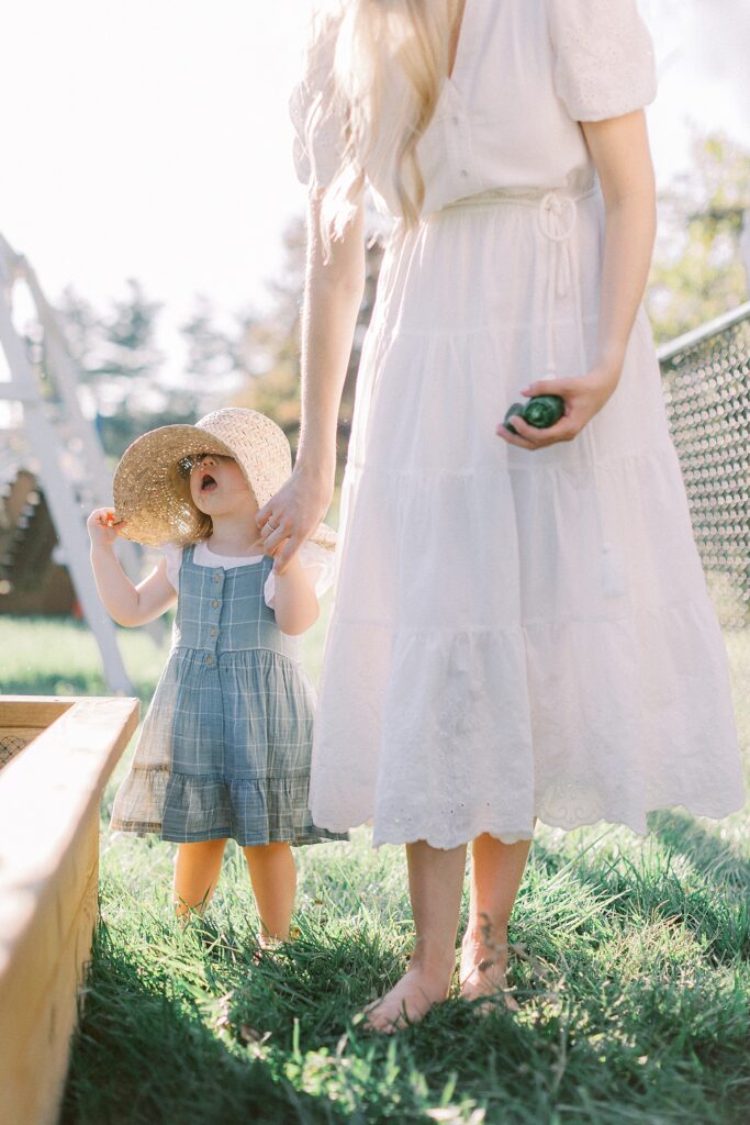 A little girl in a straw hat walks with her mother in their backyard garden in a photo by Katelyn Ng Photography.