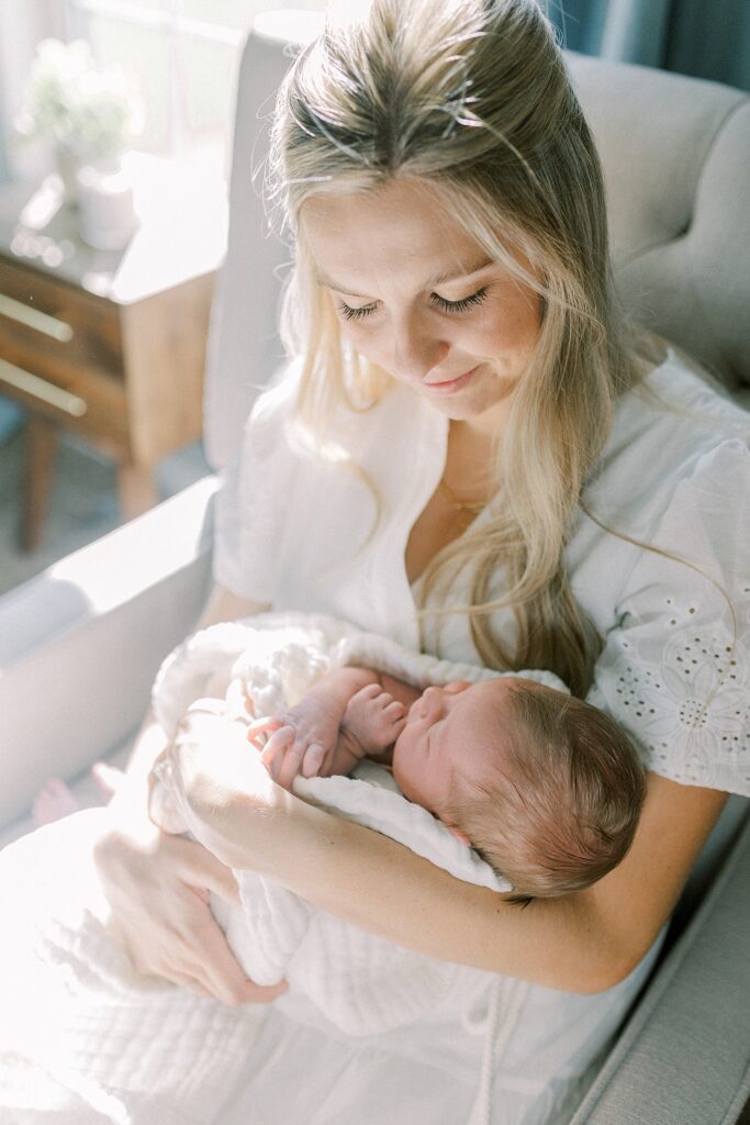 A new mother snuggles her baby boy in a rocking chair in front of a sunny window in their nursery in a portrait by Indianapolis based newborn photographer, Katelyn Ng.