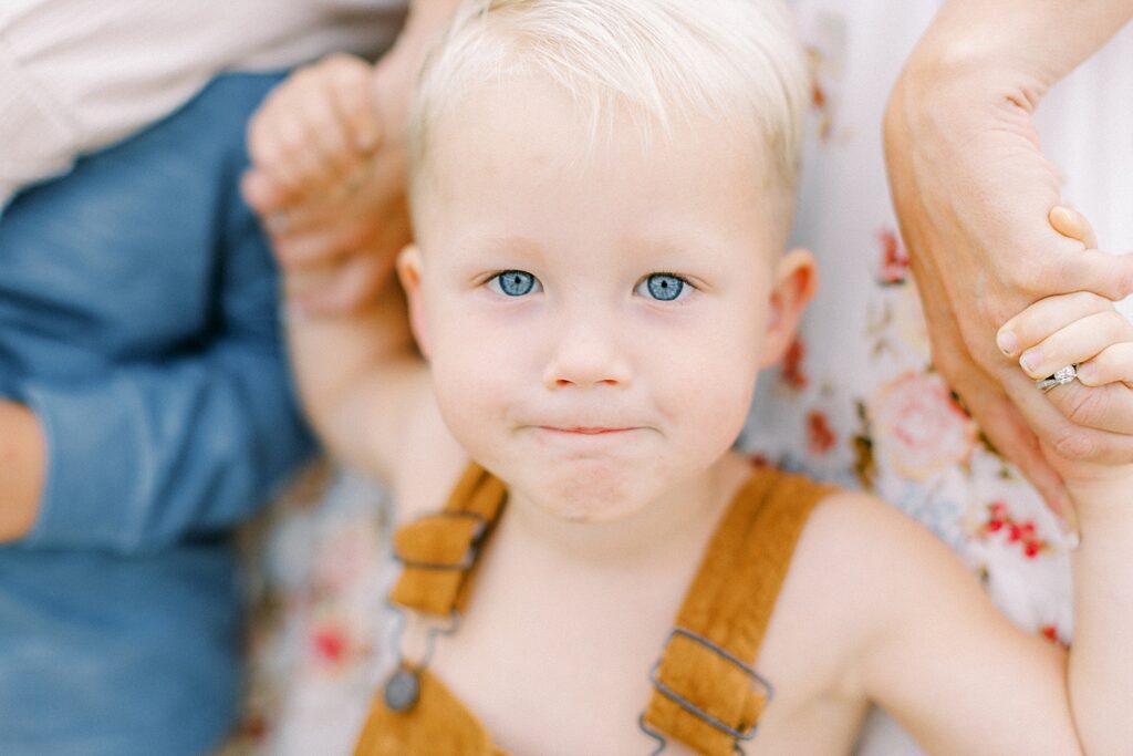 A young blonde boy with blue eyes looks into the camera while holding his mother's hands during family photos with Bloomington, Indiana photographer Katelyn Ng.