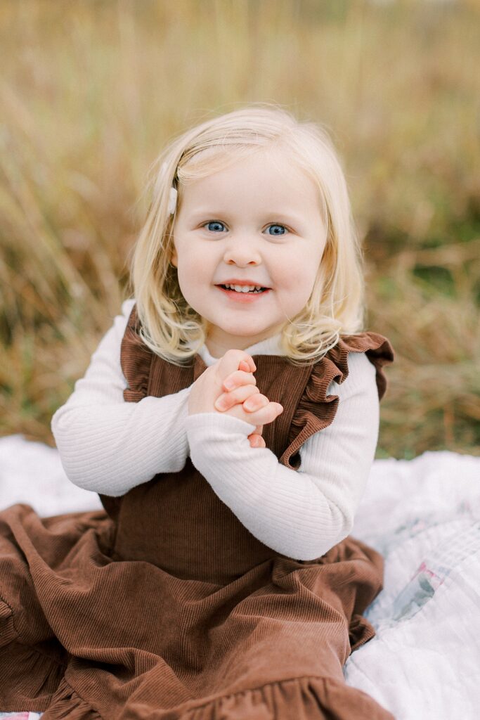 A little girl wearing a brown dress and blonde hair smiles at the camera during family photos by Carmel, Indiana photographer Katelyn Ng Photography.