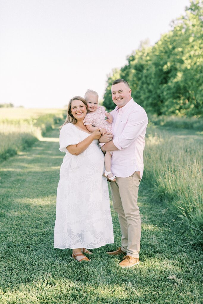 A pregnant woman wearing a white dress poses with her husband and daughter during their family photos by Indianapolis family photographer Katelyn Ng Photography.