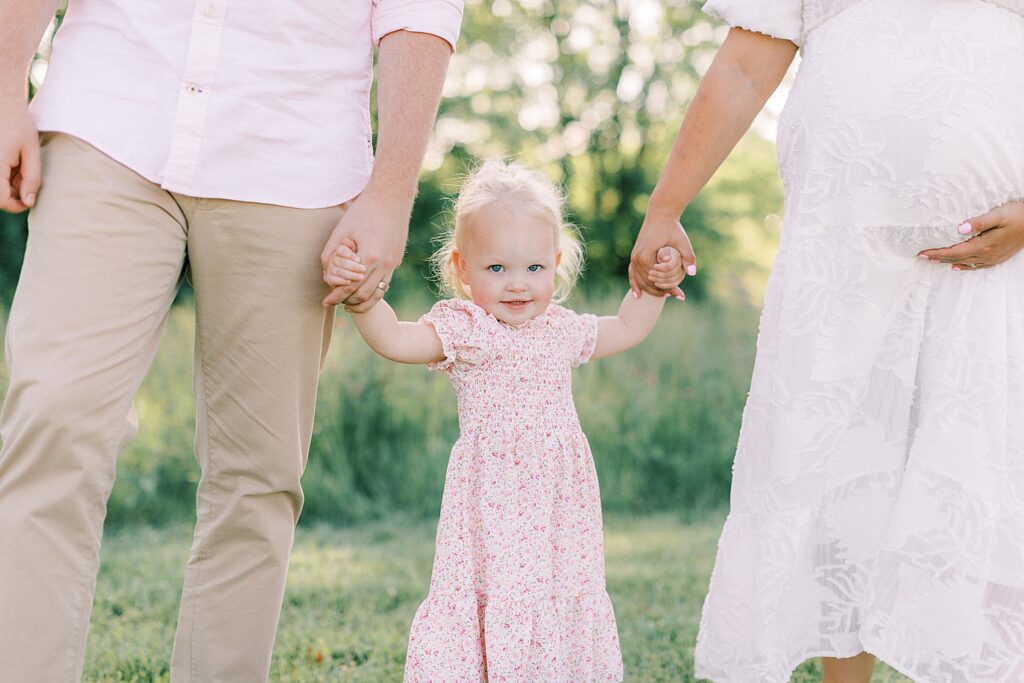 A little blonde girl in a pink floral dress holds her mother and father's hands and poses for Indianapolis family photographer Katelyn Ng Photography.