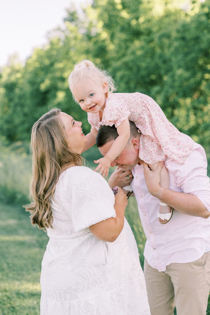 A little girl in a pink floral dress sits on her father's shoulders, leaning down to stroke her mother's face in a photo by Indianapolis family photographer Katelyn Ng Photography.