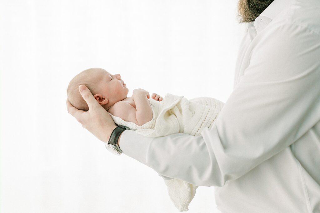 A father holds his baby girl up in front of a window as the cream blanket that surrounds her keeps her warm.