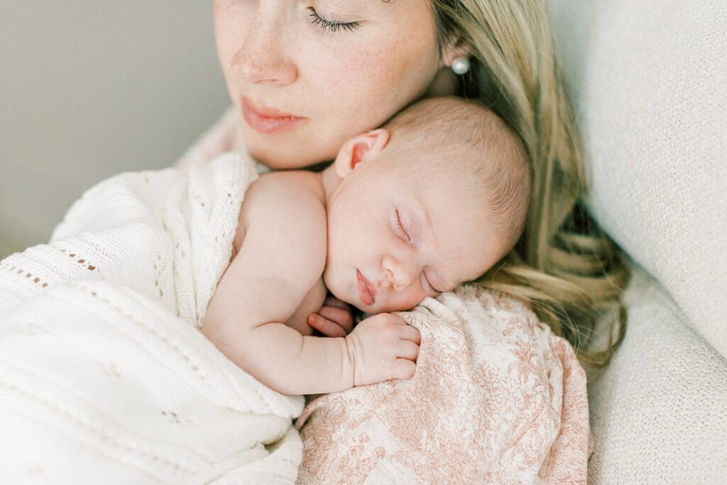 A mother wearing a toile pink and white dress closes her eyes and holds a sleeping baby on her shoulder as she rocks her to sleep in a photo by newborn photographer Katelyn Ng Photography