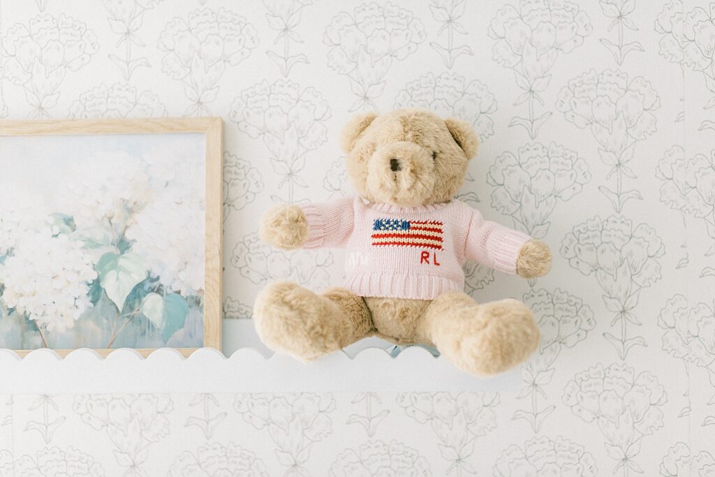 A little Ralph Lauren style teddy bear wearing a sweater perches on a shelf in a classic style nursery. 